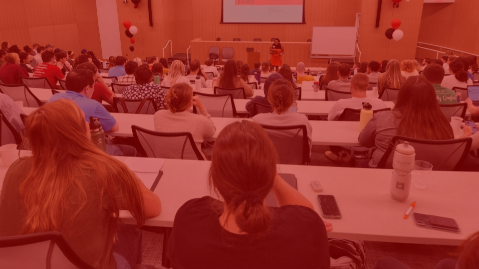 Picture of a large lecture classroom taken from the back of the room. The backs of students heads are visible and someone is speaking at the front of the class as a podium. 